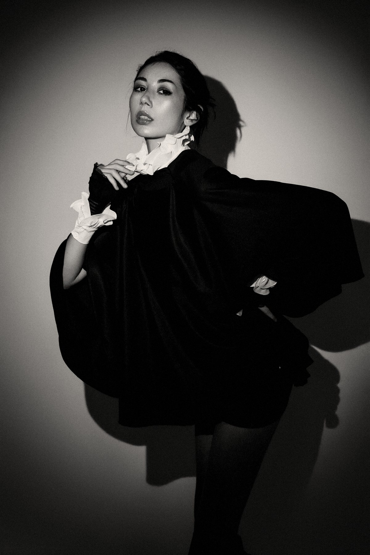 Duda com vestido preto volumoso e punhos brancos, projetando sombra em uma parede clara.